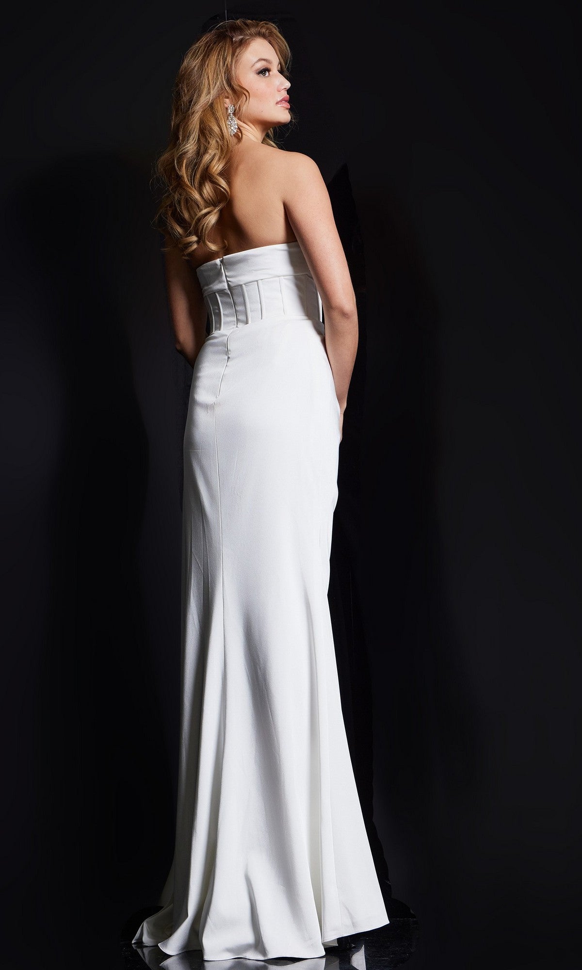 A woman with long wavy hair models the Jovani 36988 long prom dress featuring a strapless sweetheart neckline and fitted corset bodice, standing against a black background and facing away from the camera.